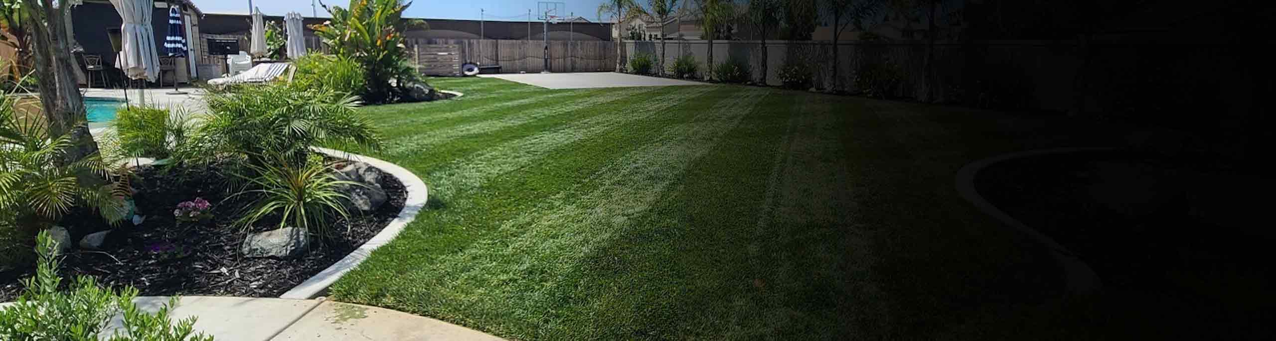A scenic backyard with concrete sidewalk, concrete landscape border and concrete basketball court alongside green grass.